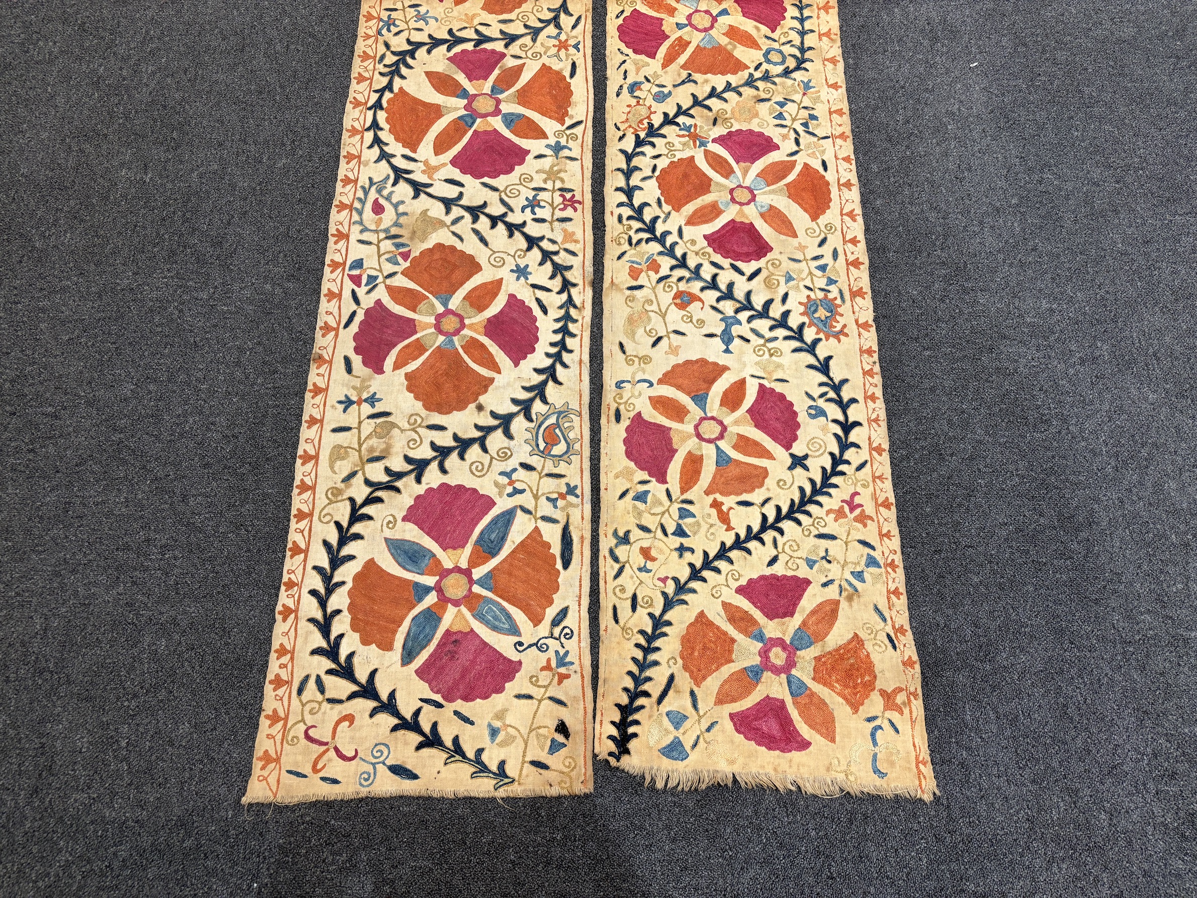 A pair of Uzbekistan long wide panels, both embroidered in mostly red, coral and blue with large symbolic flowers and trailing vines on woven cotton using mostly Burkara couching, longest 250cm long x 38cm wide